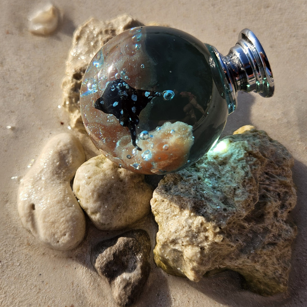A small, round resin cabinet knob er lies on sand among smooth and rough rocks.
